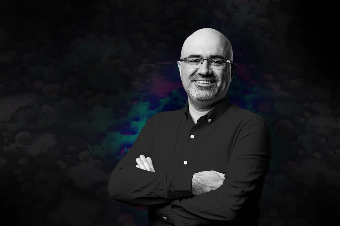 Confident man with glasses and beard smiles with arms crossed against a dark abstract background, conveying expertise and approachability.