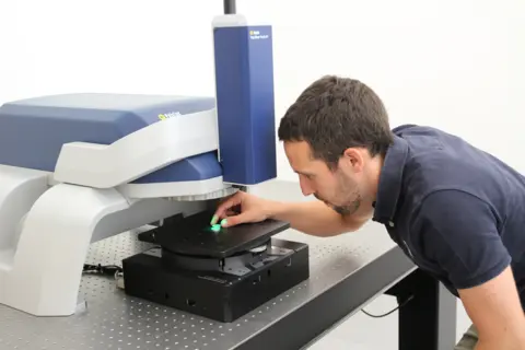Engineer calibrating a blue and white Polytec TopMap Pro.Surf optical profiler on a precision metal component on a lab table.