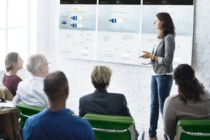 Businesswoman giving a professional presentation using a digital whiteboard to a seated diverse audience in a bright office.