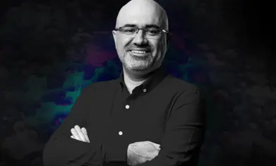 Confident man with glasses and folded arms smiling against a dark, textured background, conveying professionalism and approachability.