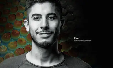 Young male service engineer Okan with short hair and beard, smiling confidently against a dark, honeycomb-patterned background.
