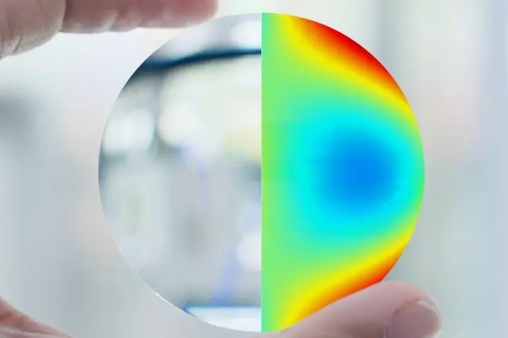 Hand holding a transparent optical lens with a colorful 3D surface topography map for precision measurement.