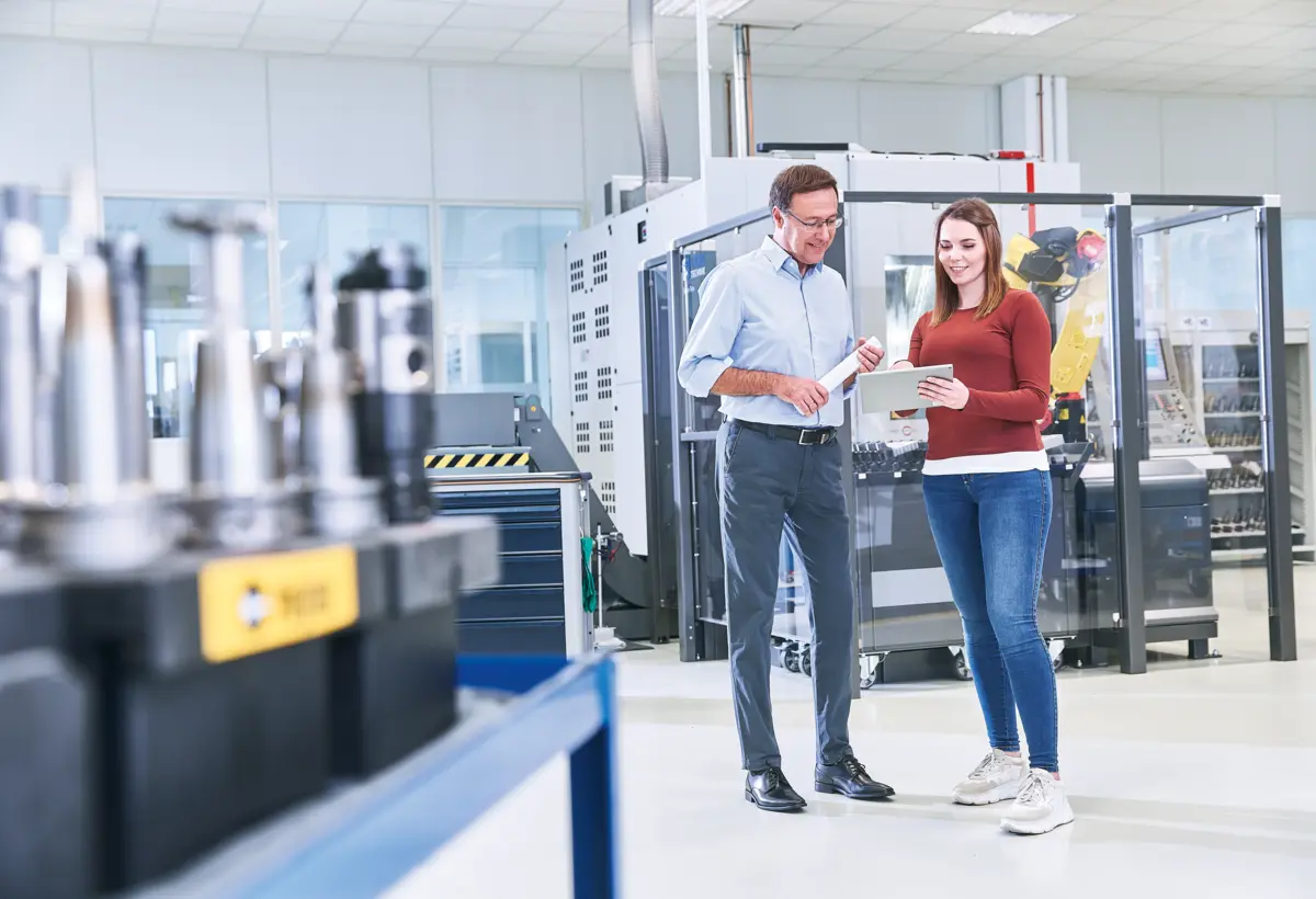 Two industrial engineers discussing data on a tablet in a modern manufacturing facility with advanced machinery.