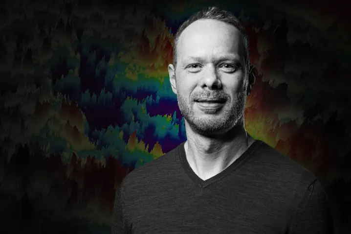 Man with a confident expression standing against a dark background with colorful surface profiling data visualization.
