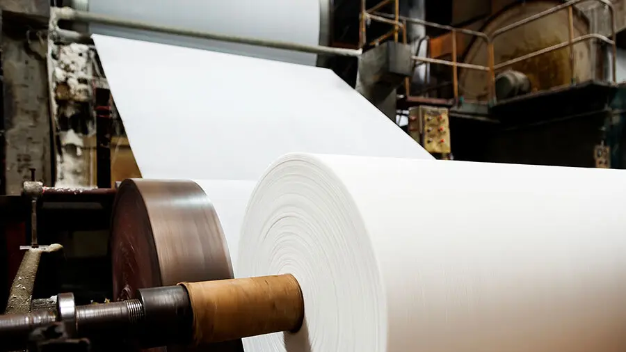Large roll of white paper being processed on an industrial machine in a manufacturing facility