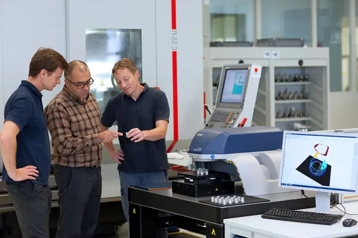 Three engineers discuss results next to a Polytec 3D optical surface profiler displaying measurement data on a monitor.